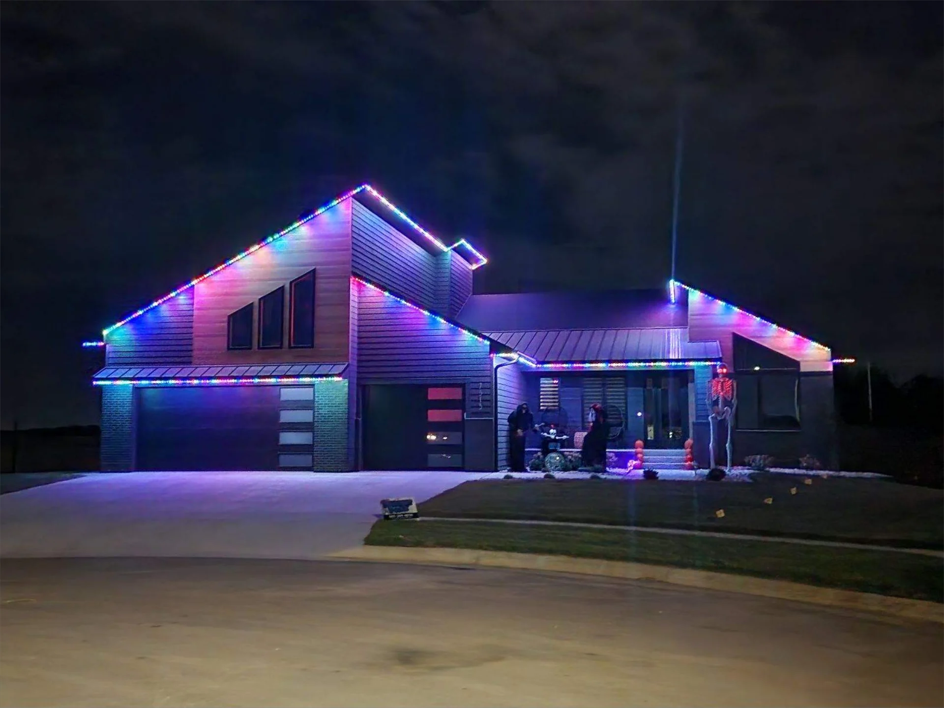 House decorated with purple and blue holiday lights at night, with a lit front porch and driveway.