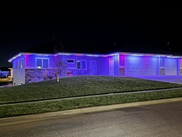 Single-story house at night illuminated with blue and purple exterior lights behind a grassy lawn.
