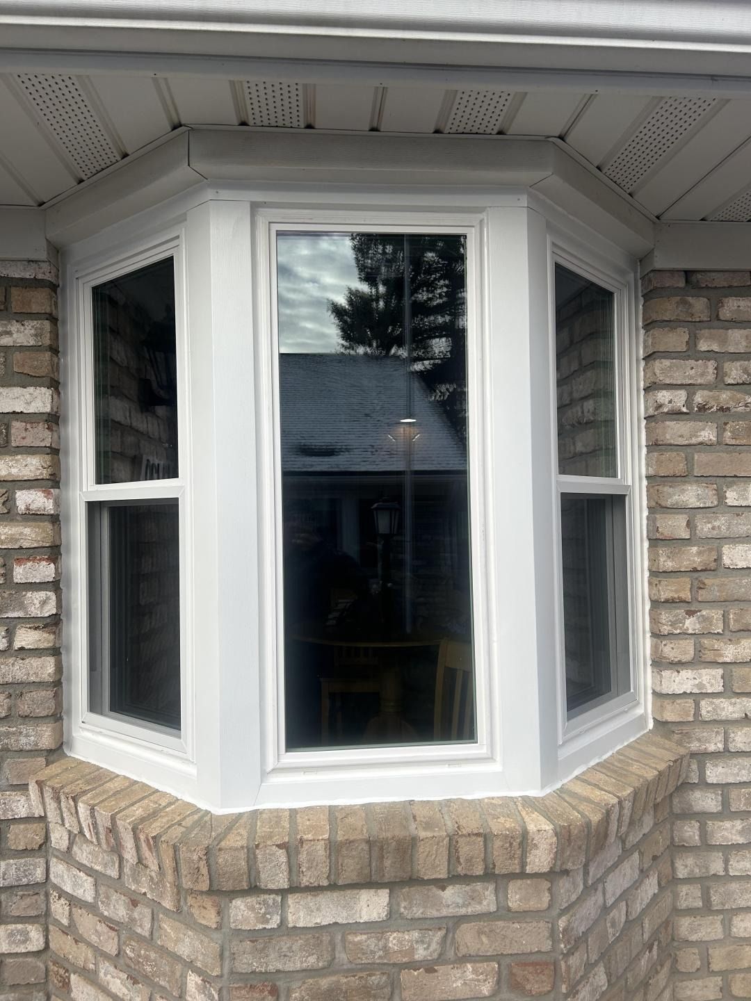 White bay window set in a tan brick house wall under the roof overhang.