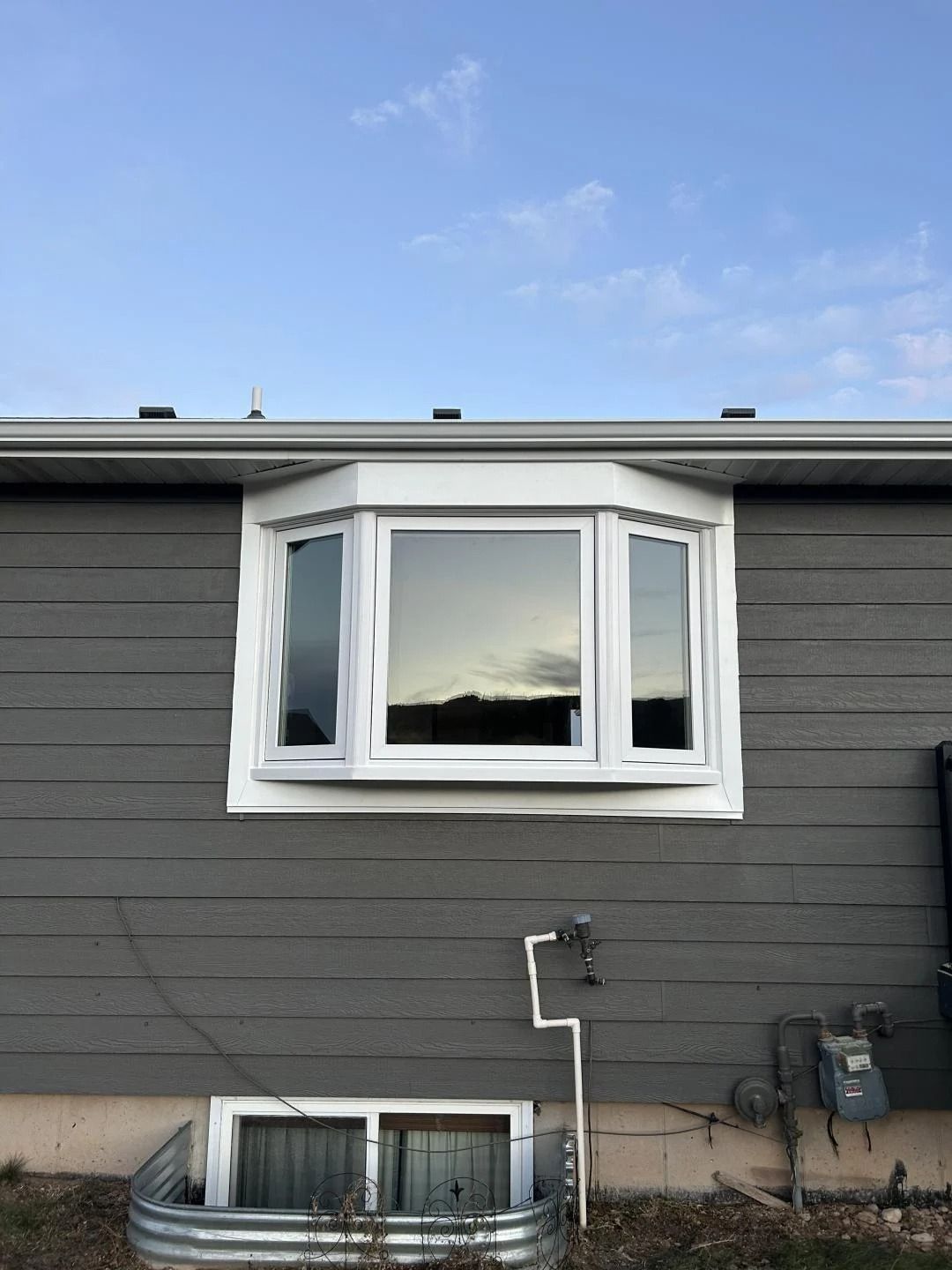 Gray house exterior with white bay window and basement window beneath a blue sky
