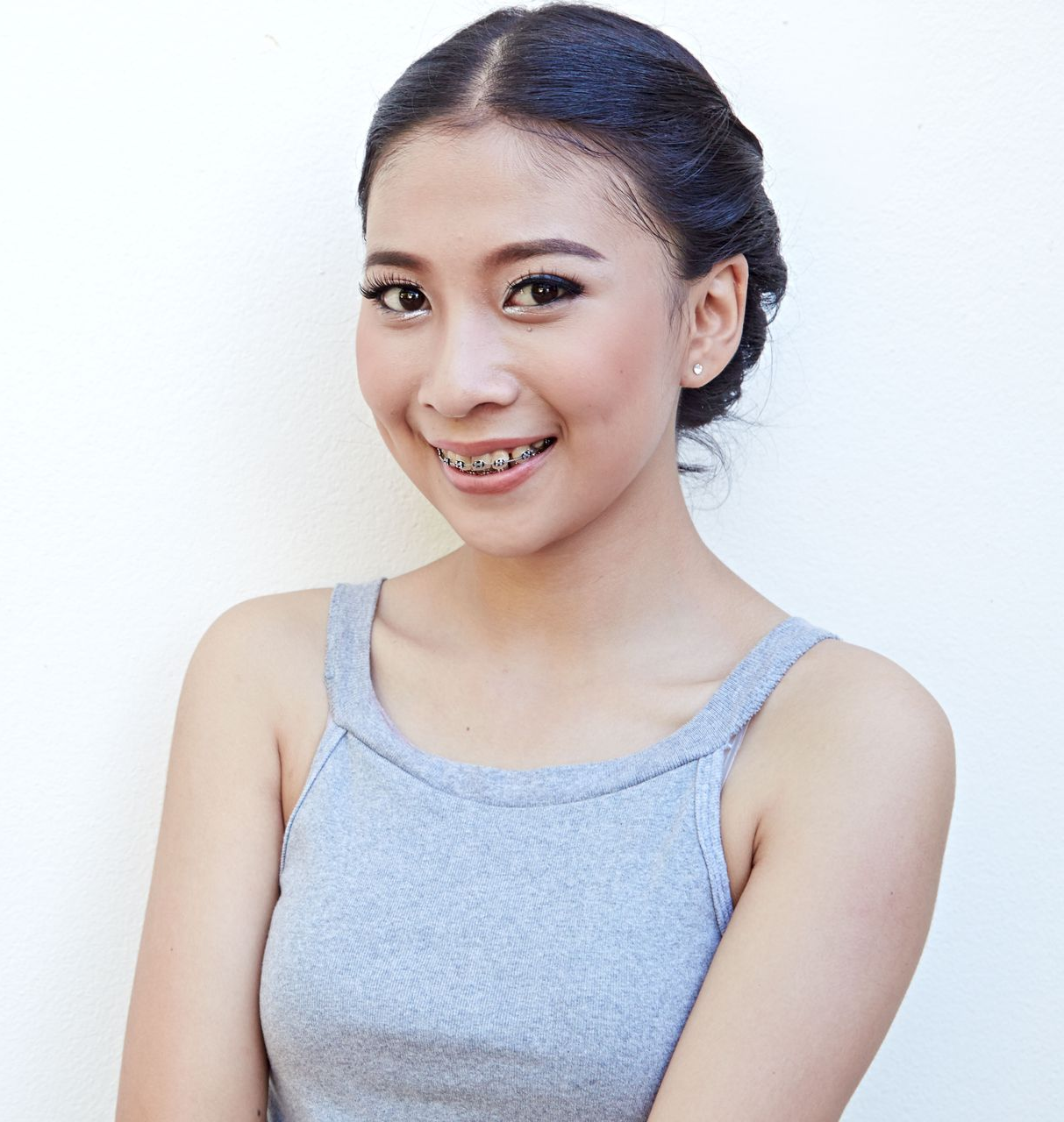 Woman smiling, wearing braces and a gray tank top, against a white wall.