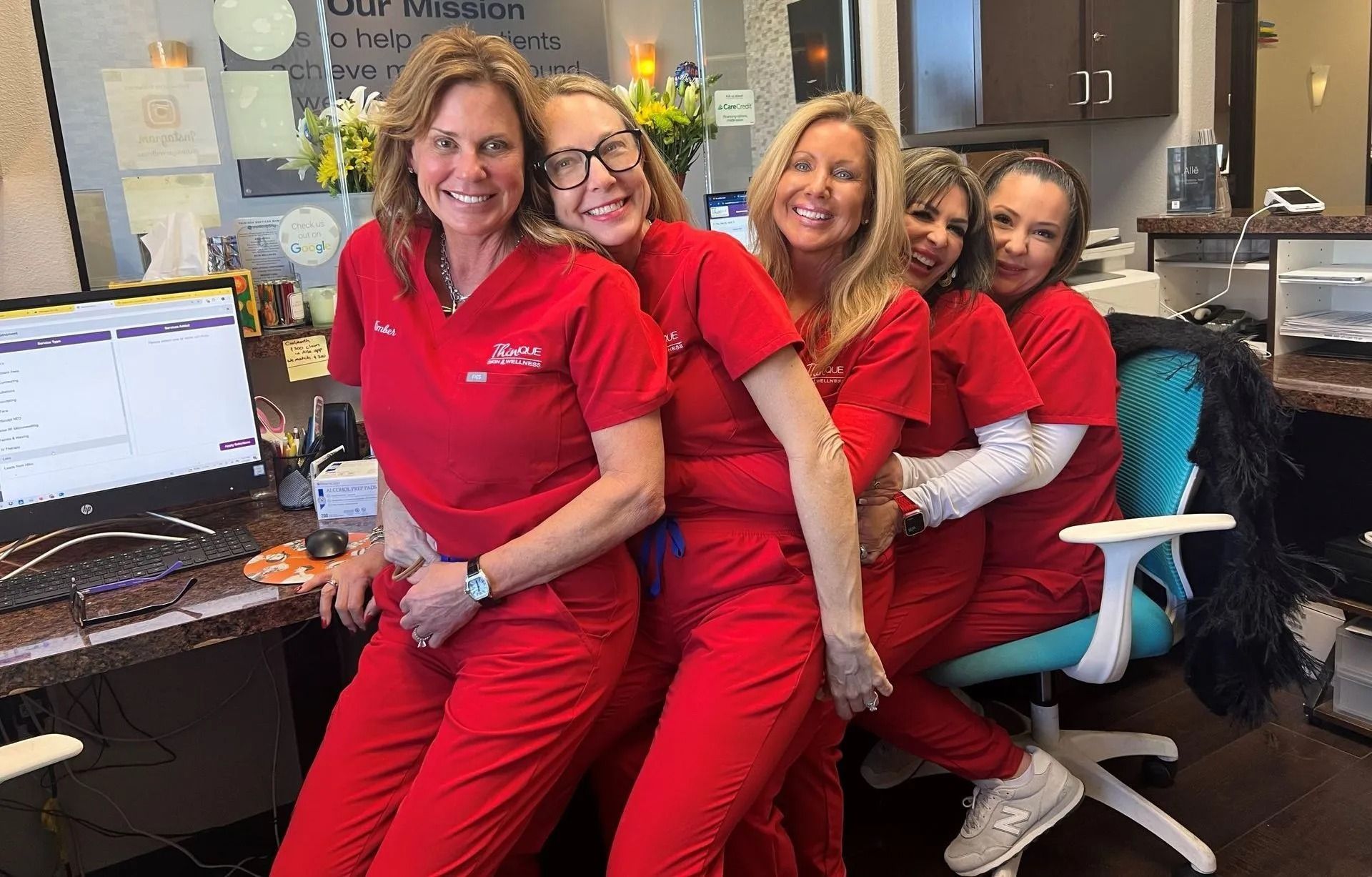 Five smiling people in red scrubs, posing together at a desk in an office.