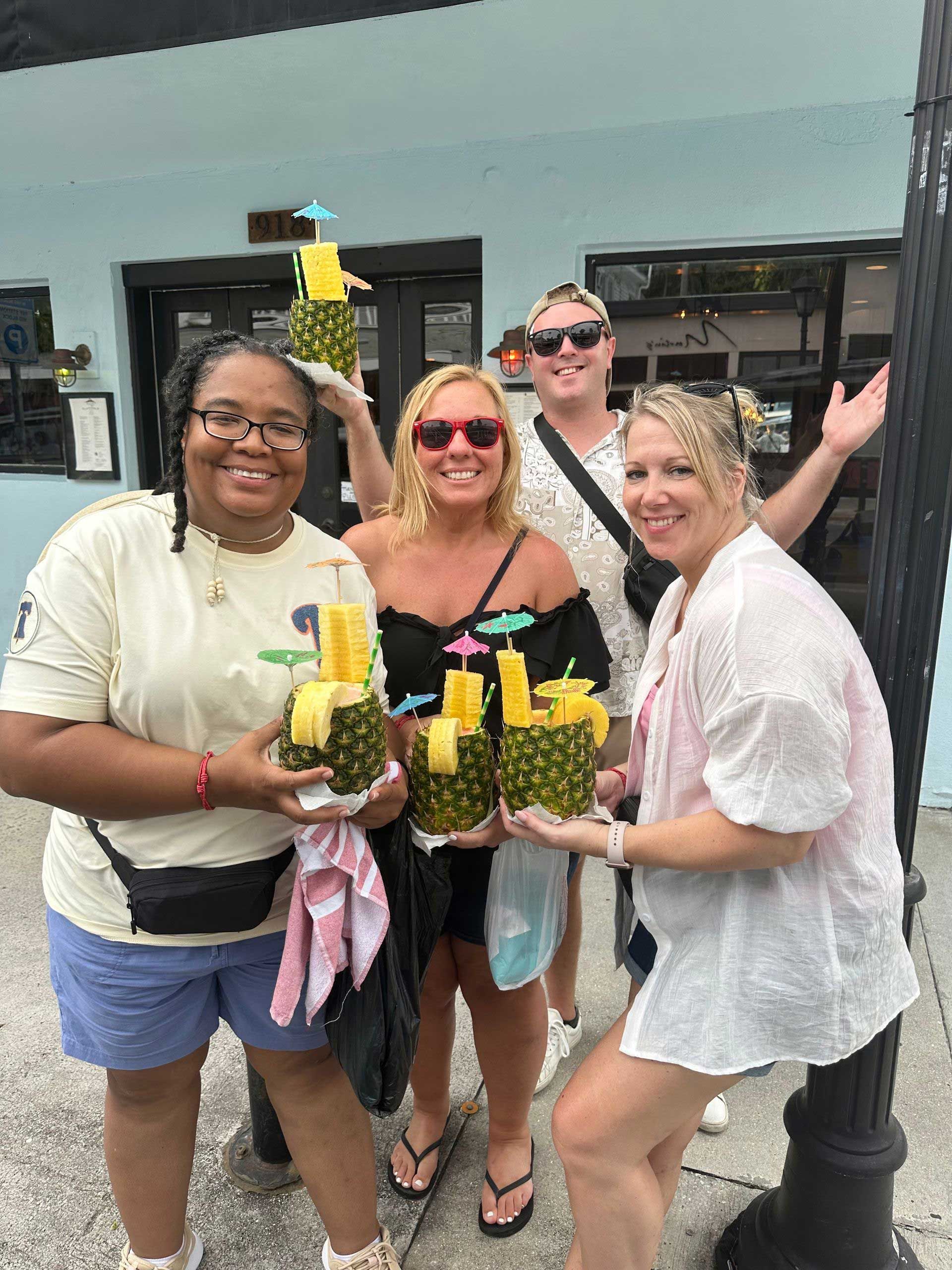four people smiling, holding pineapple drinks outside a building