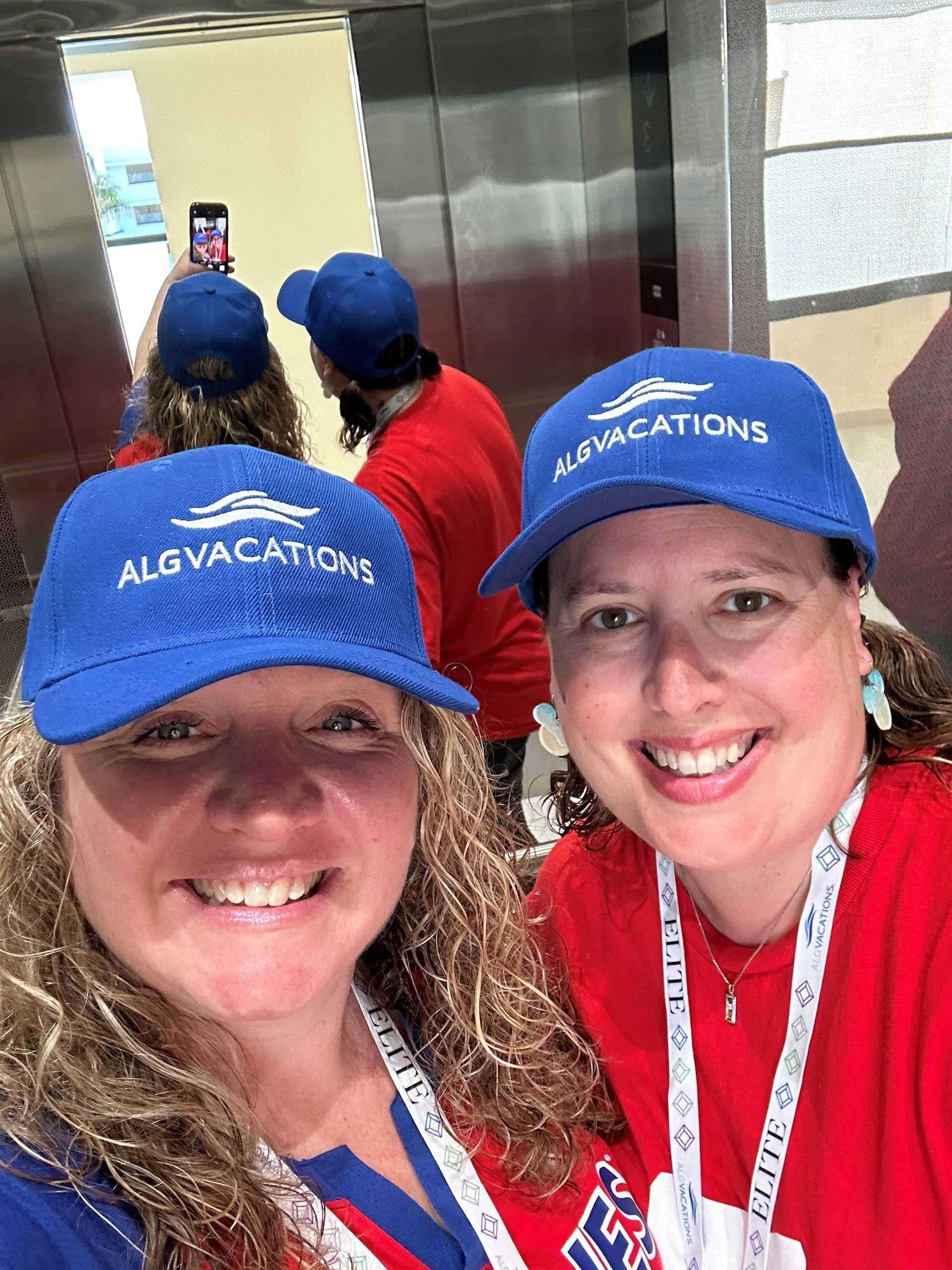 two women in matching blue hats and red shirts smiling inside an elevator, taking a selfie