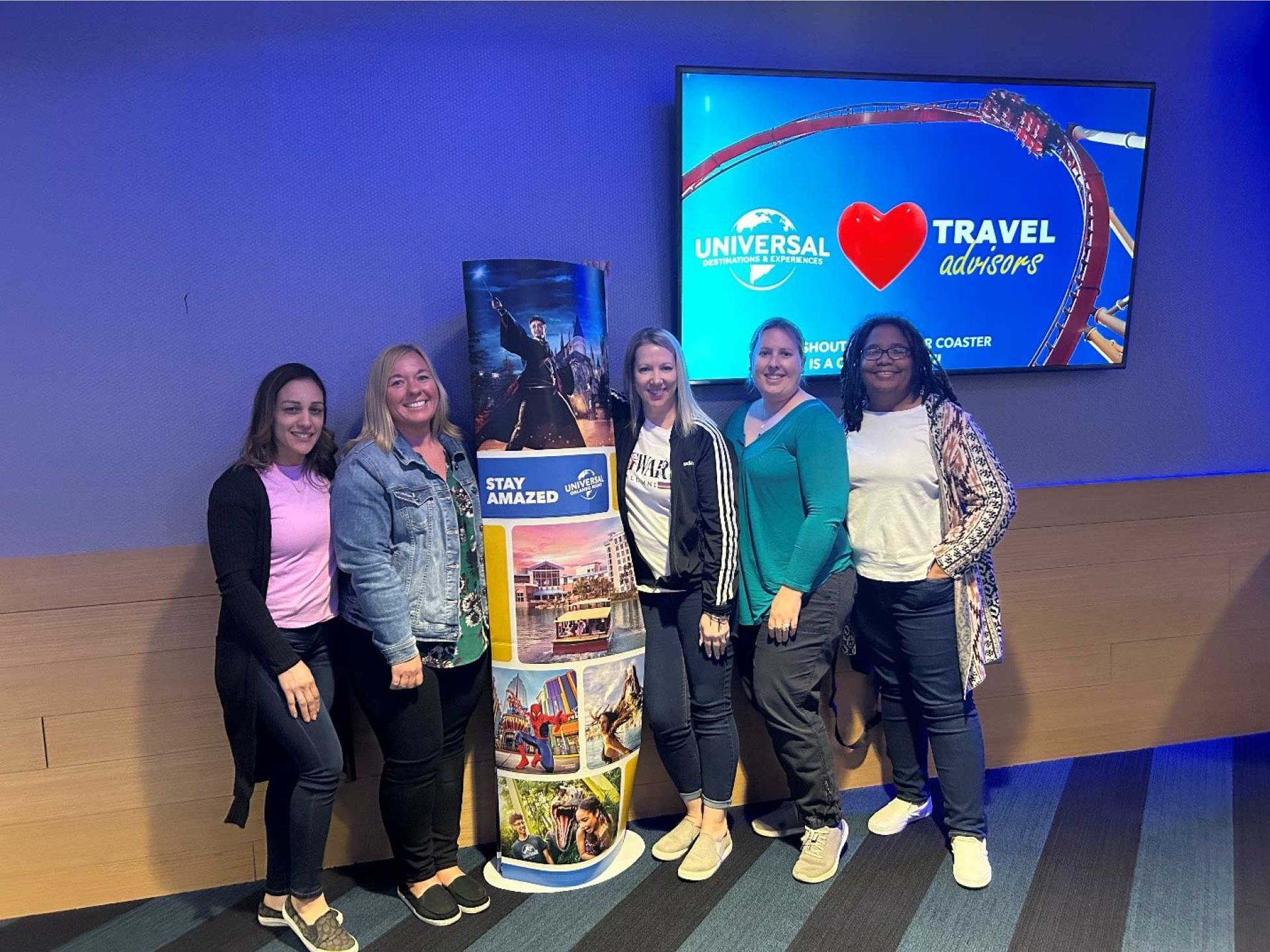 five women pose in front of a TV screen and promotional display