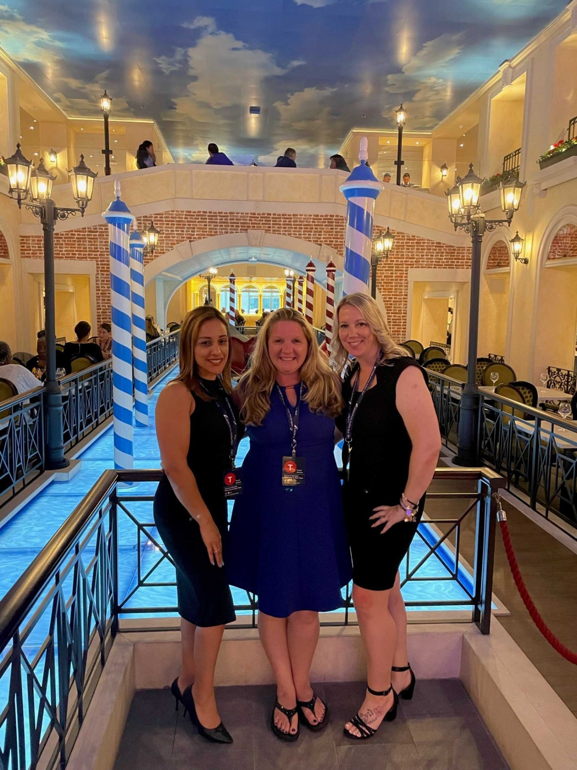 three women smiling and posing together in a Venetian-themed dining area, with a blue sky ceiling