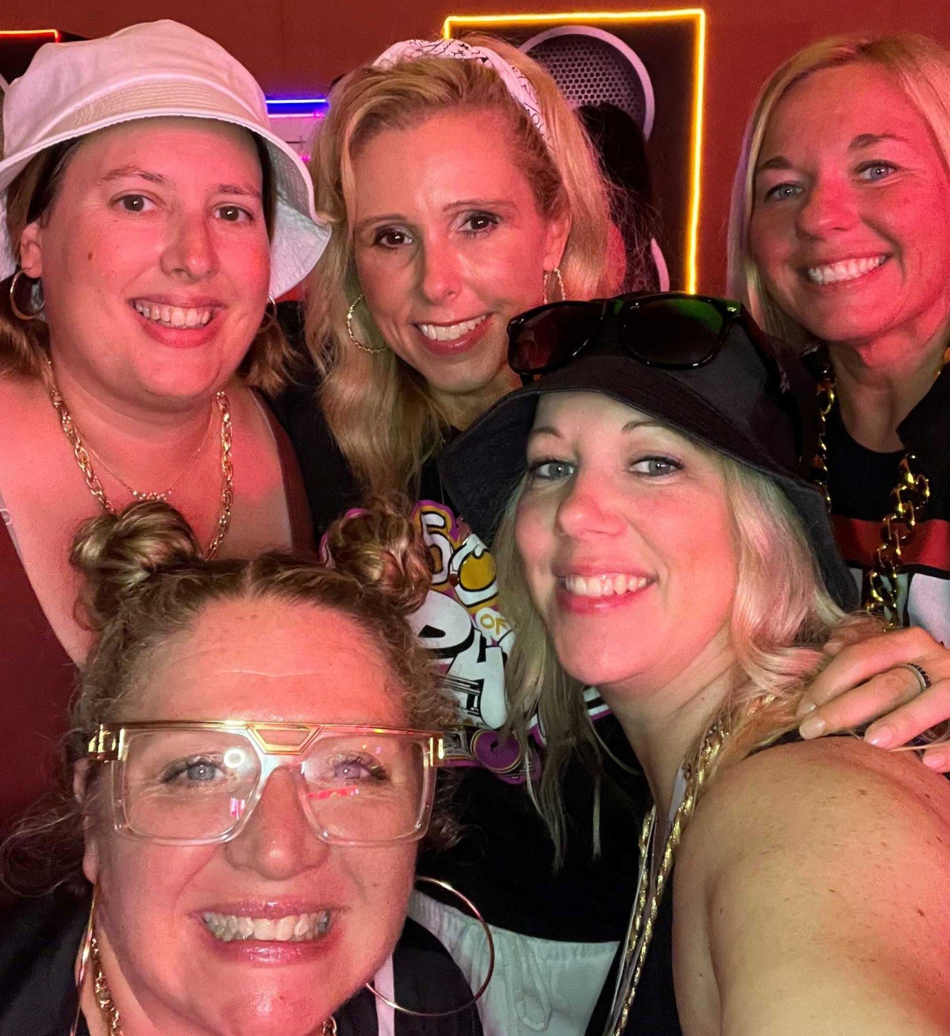 five women smiling at the camera, dressed in 80s or 90s-themed costumes with gold chains, hats, and large glasses, at a party