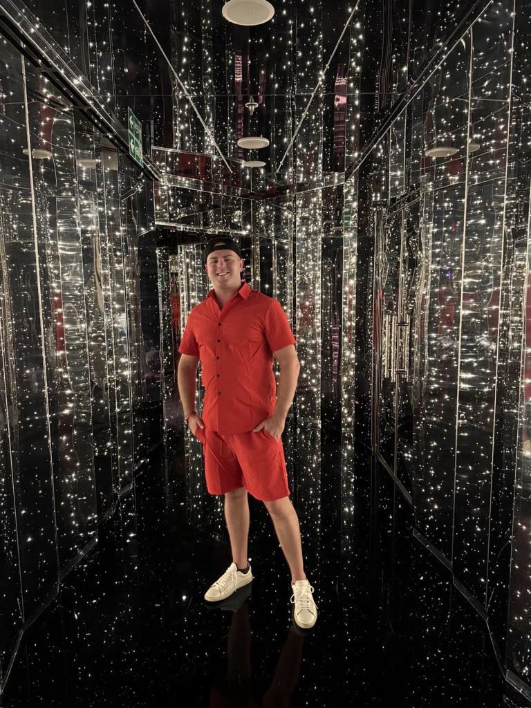 A man in a red shirt and shorts is standing in a mirrored room.