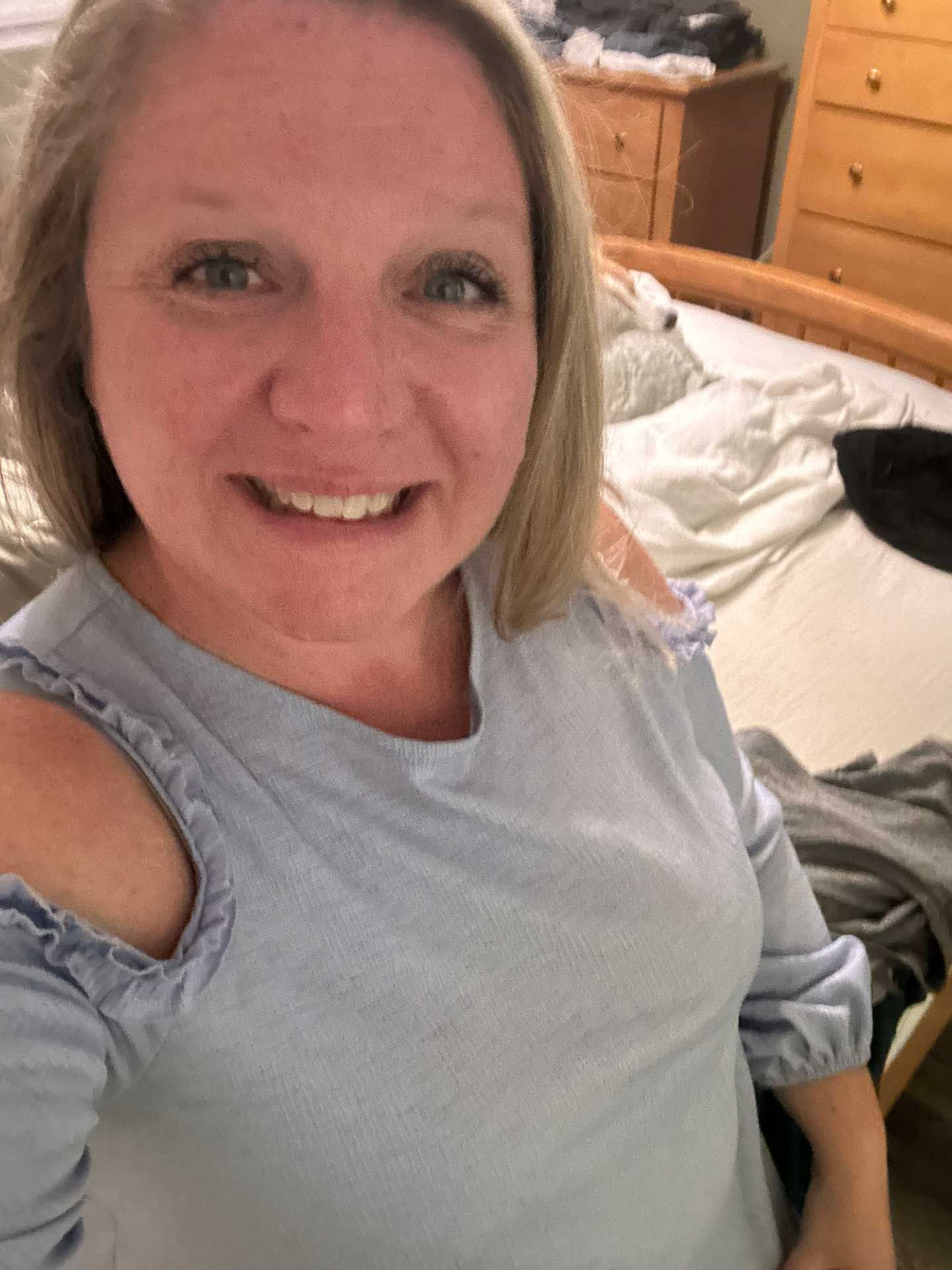 woman with blonde hair smiles, wearing a top with cut-out shoulders, in a bedroom