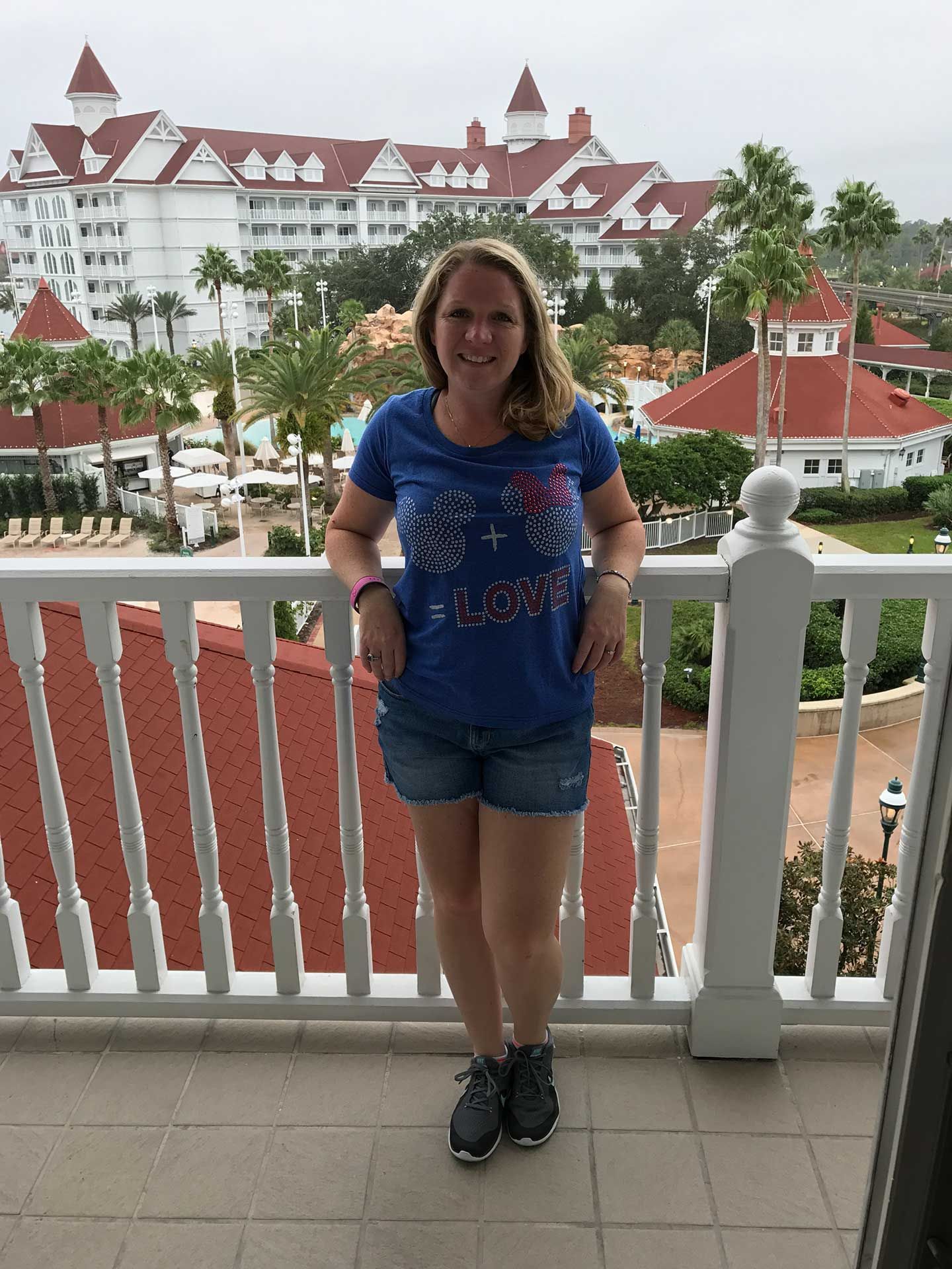 a woman in blue shirt and denim shorts stands on a balcony overlooking a resort with white buildings and palm trees