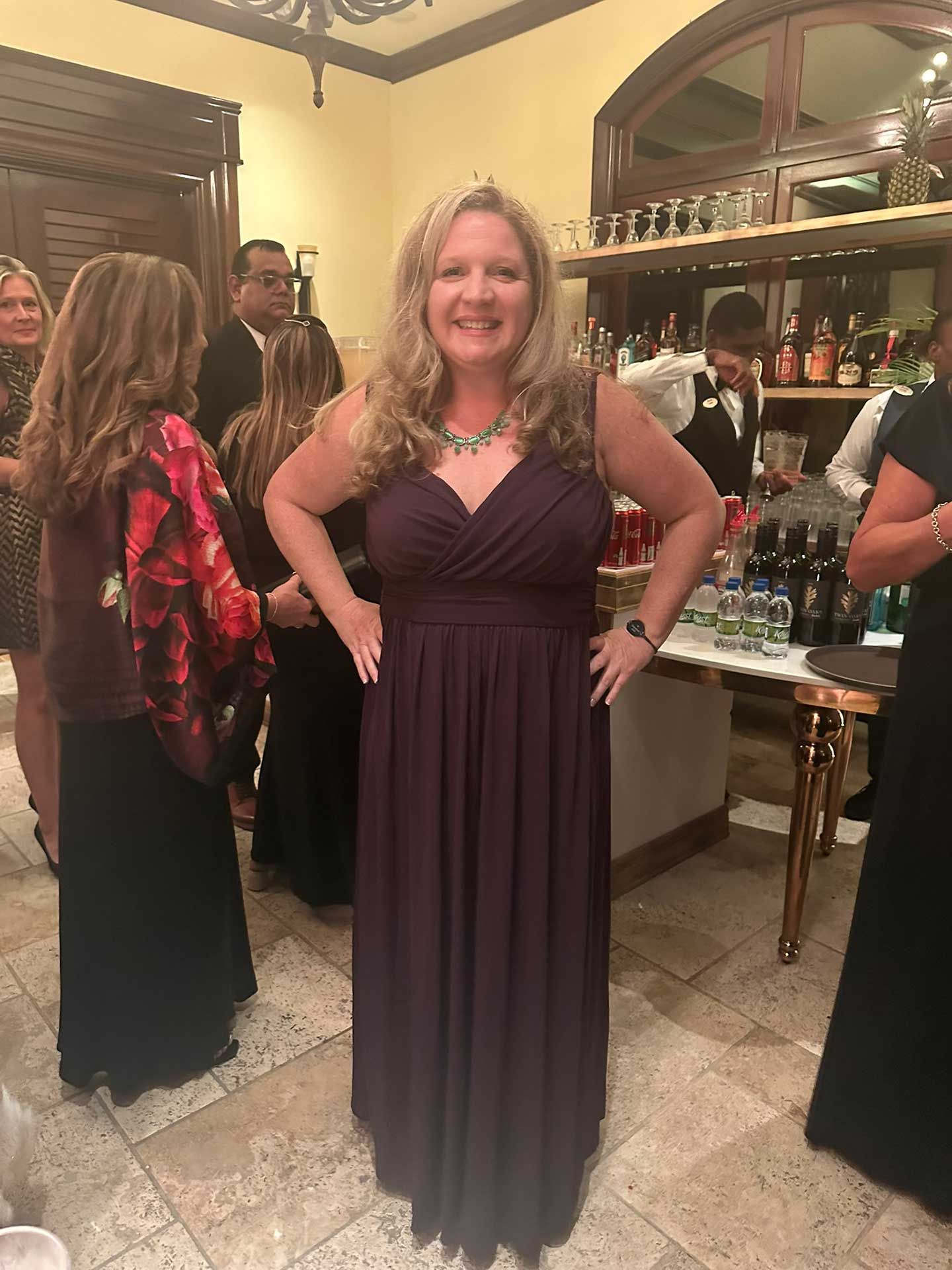 a woman in a dark purple gown smiles, hands on hips, at a formal event