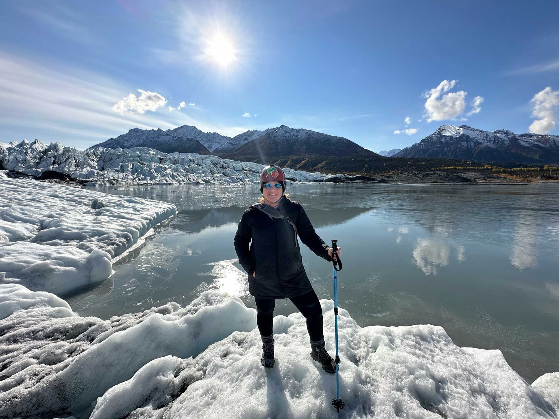 a woman hiking on ice near a glacier and mountains, under a bright sun