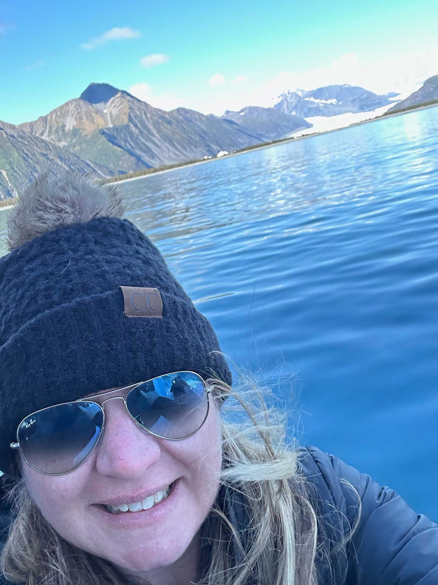 a woman in a black beanie and sunglasses smiles at the camera, mountains and water in the background