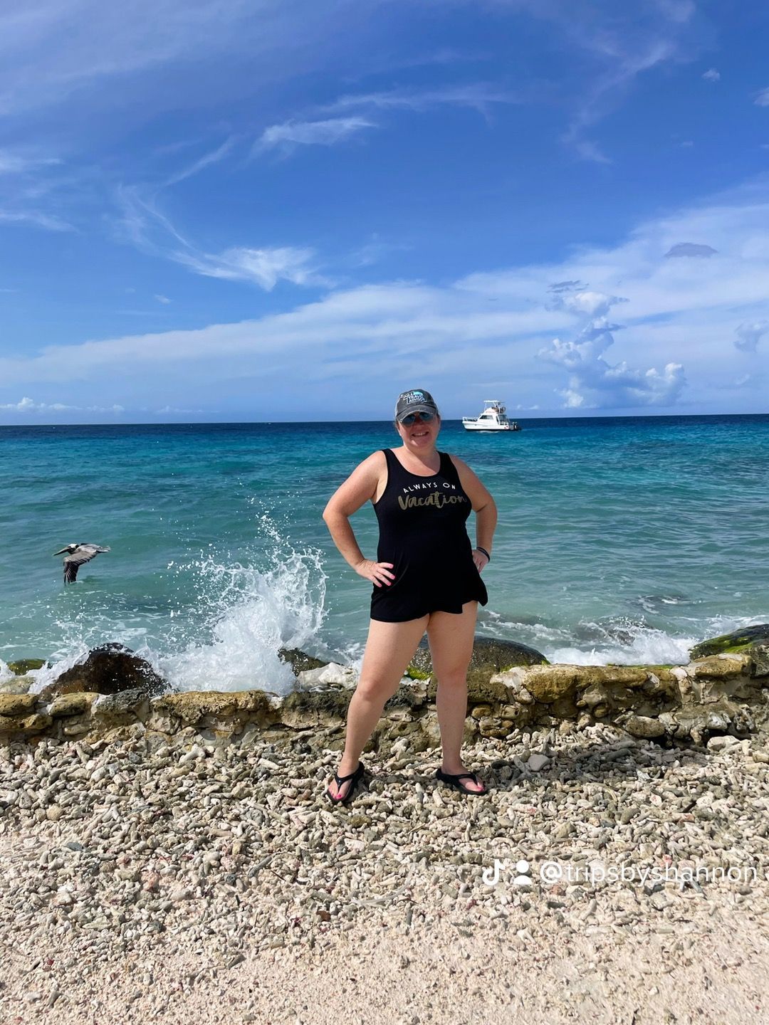a woman in black swimwear poses on a pebble beach with ocean waves crashing behind her
