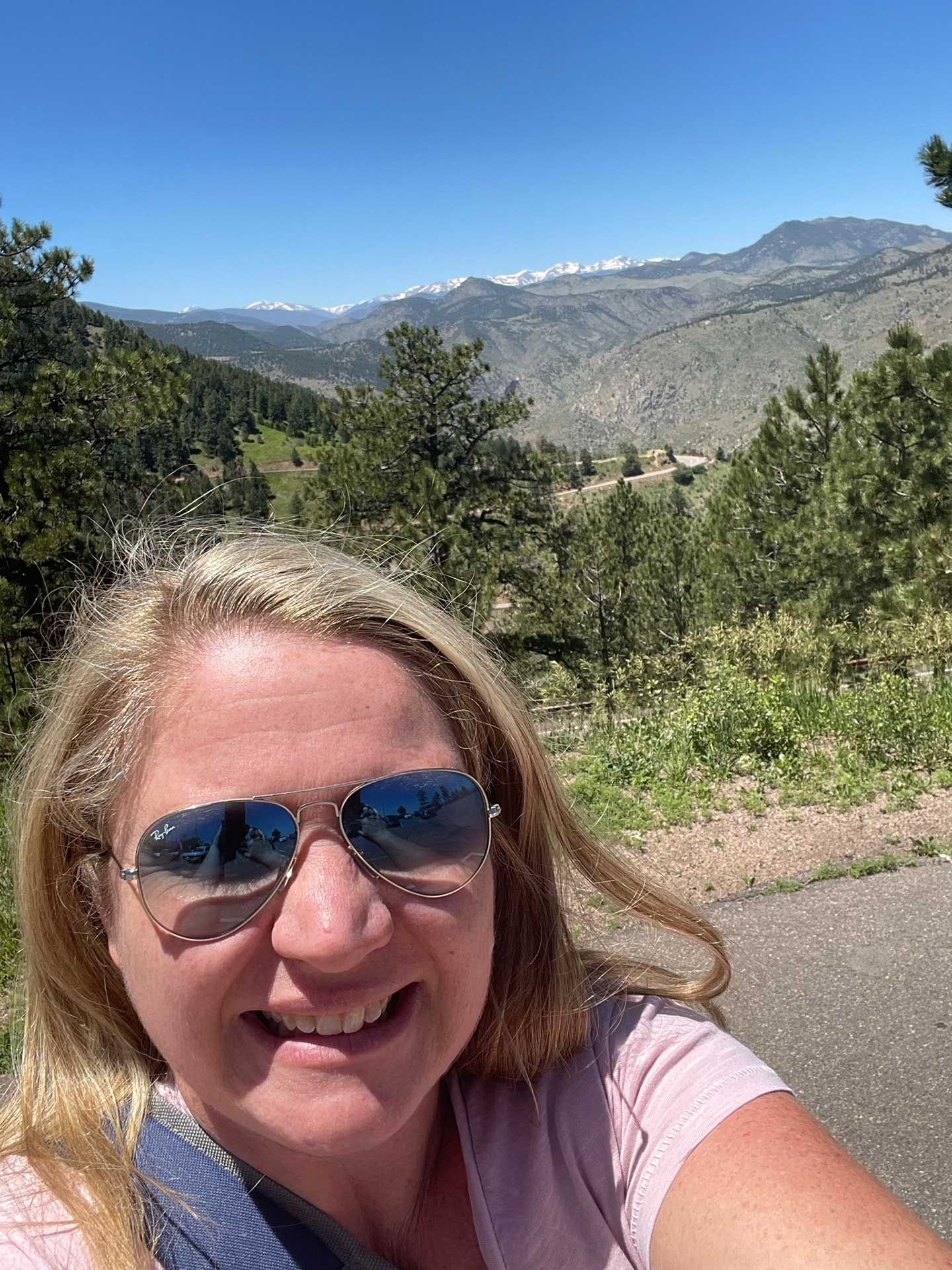 a woman in sunglasses smiles at camera outdoors, with a mountain backdrop on a sunny day