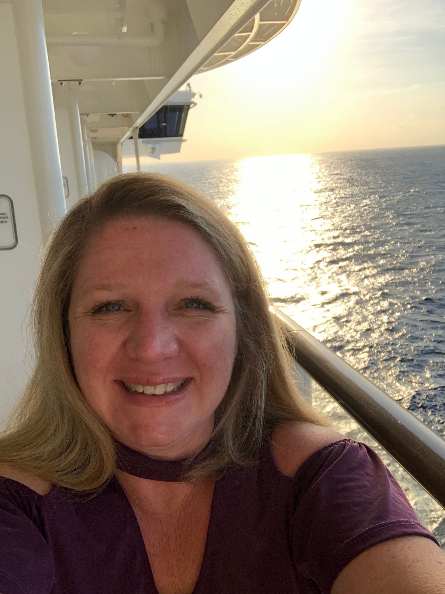 a woman smiling on a cruise ship deck at sunset, with ocean in the background