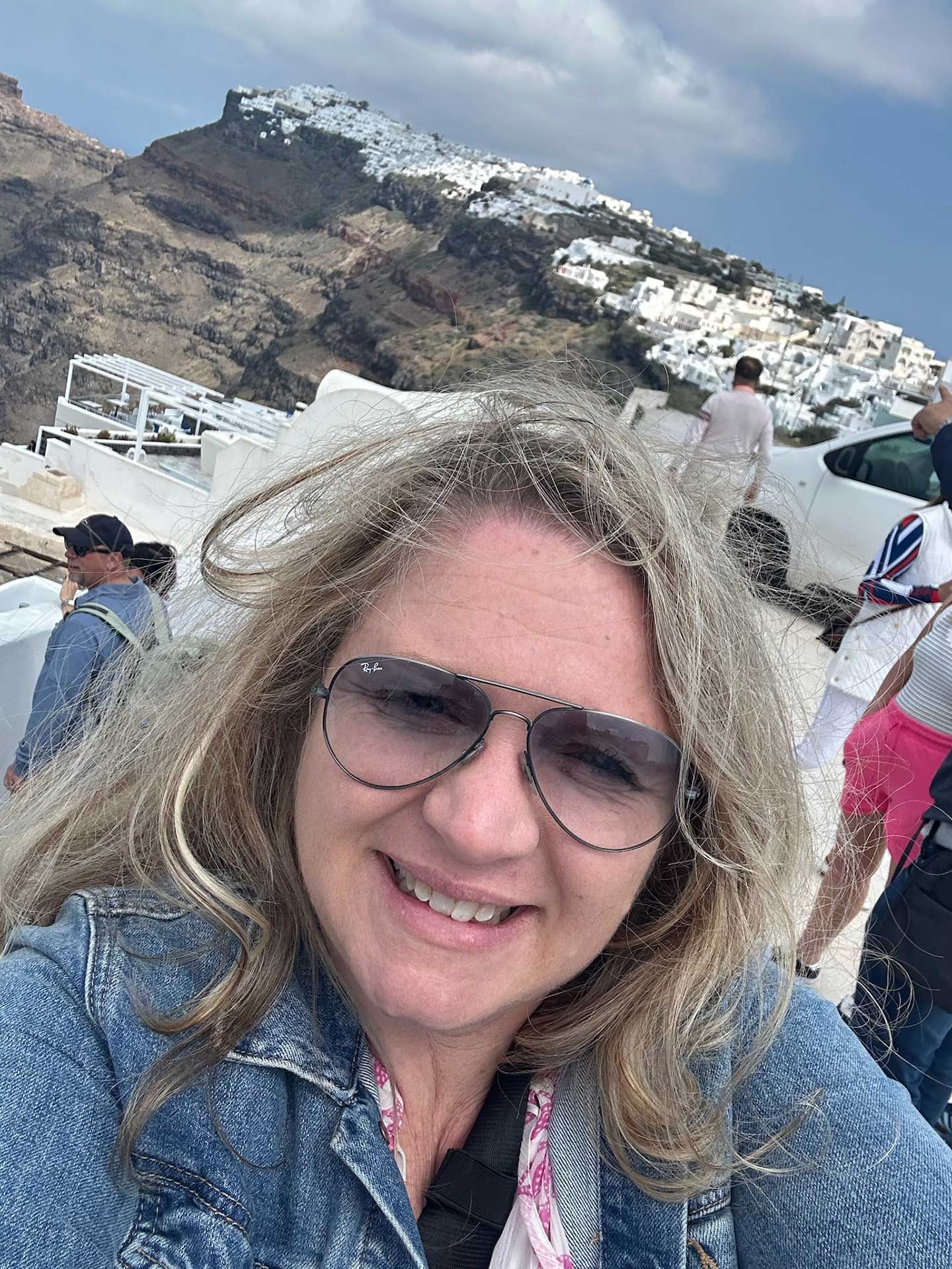 a woman in sunglasses smiles at camera, with white buildings and a mountain backdrop