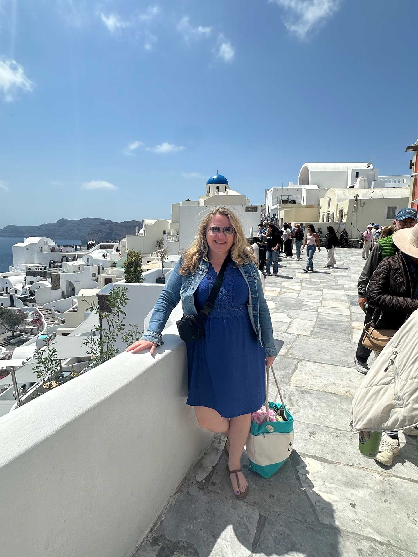 a woman in a blue dress and denim jacket, standing on a white wall overlooking the Greek island of Santorini