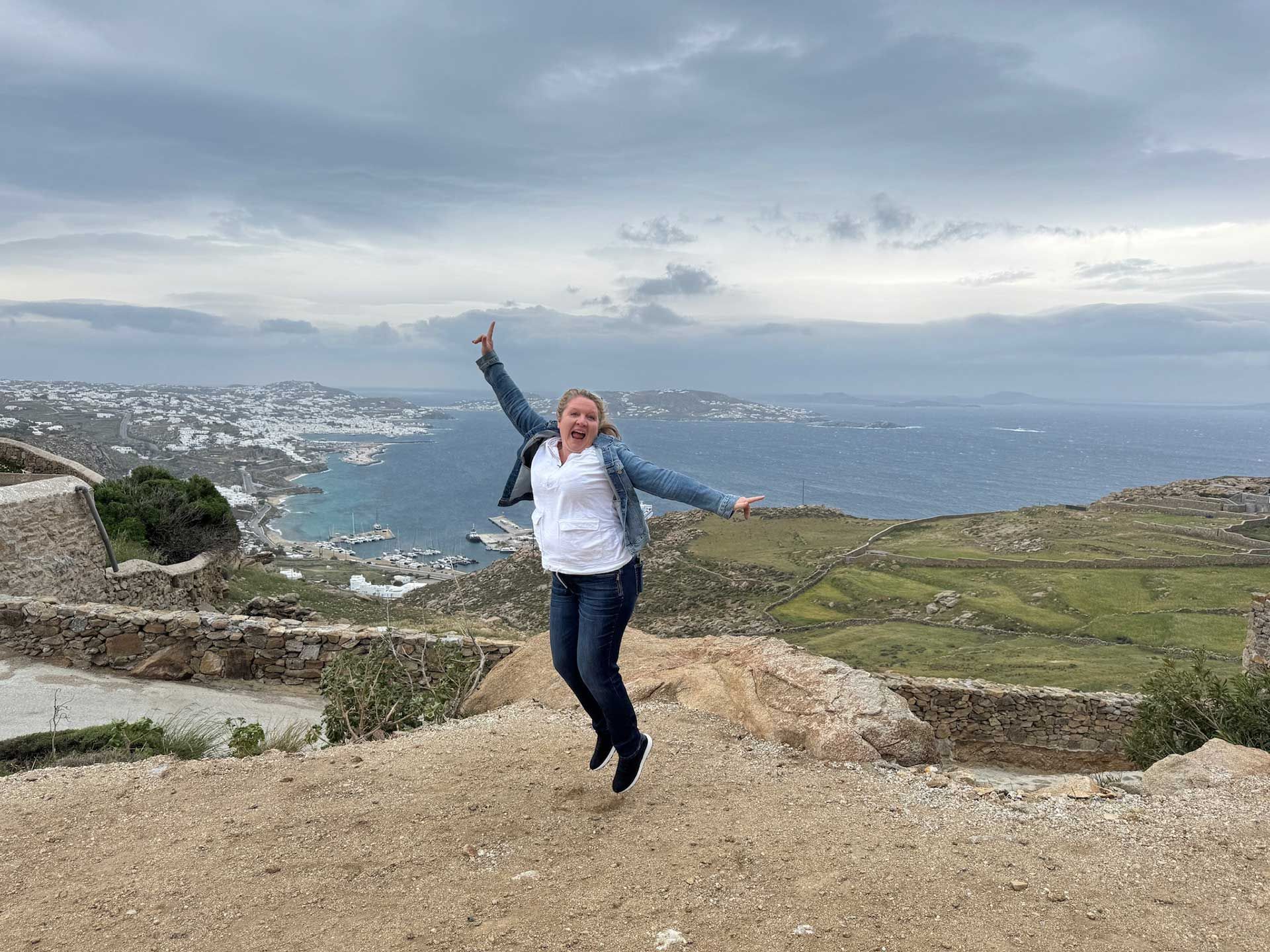 a woman jumping joyfully on a hilltop overlooking a coastal town and the sea under an overcast sky