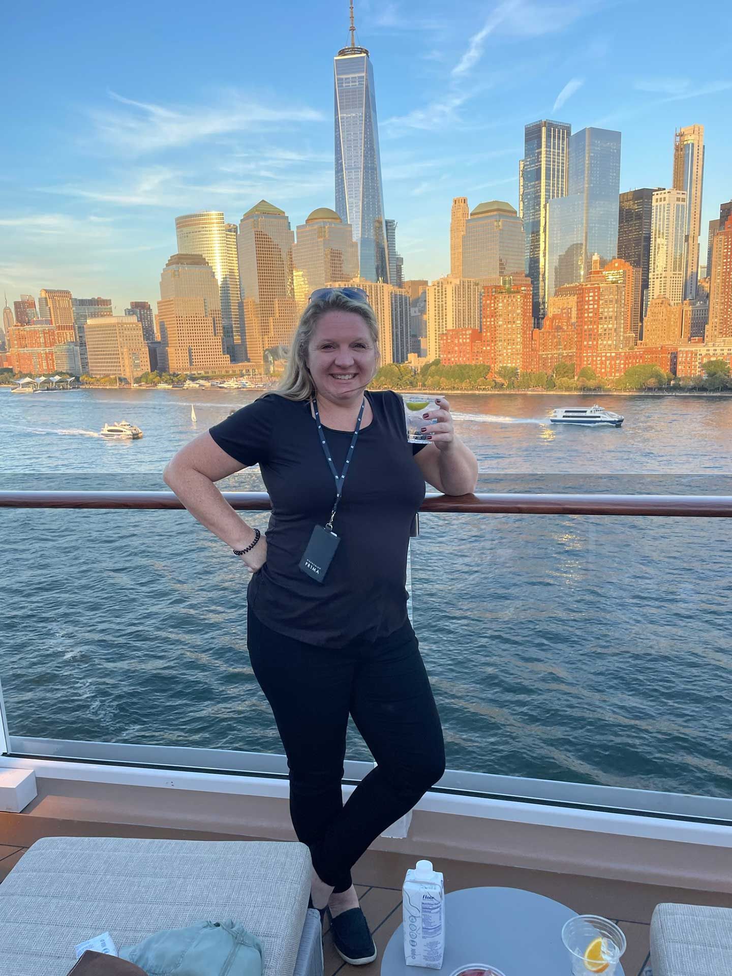 a woman on a boat deck with New York City skyline in the background, holding a drink