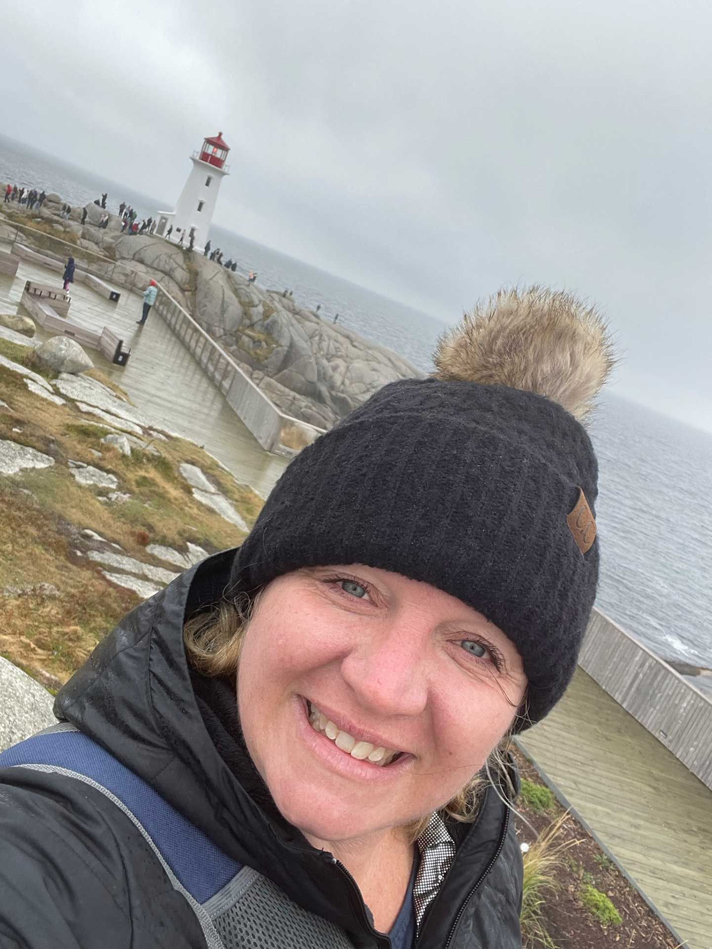 a woman in a black hat smiles at camera in front of Peggy's Cove Lighthouse on a cloudy day