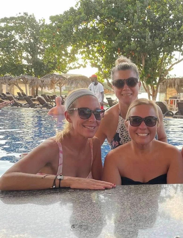 Three women wearing sunglasses are standing next to a swimming pool