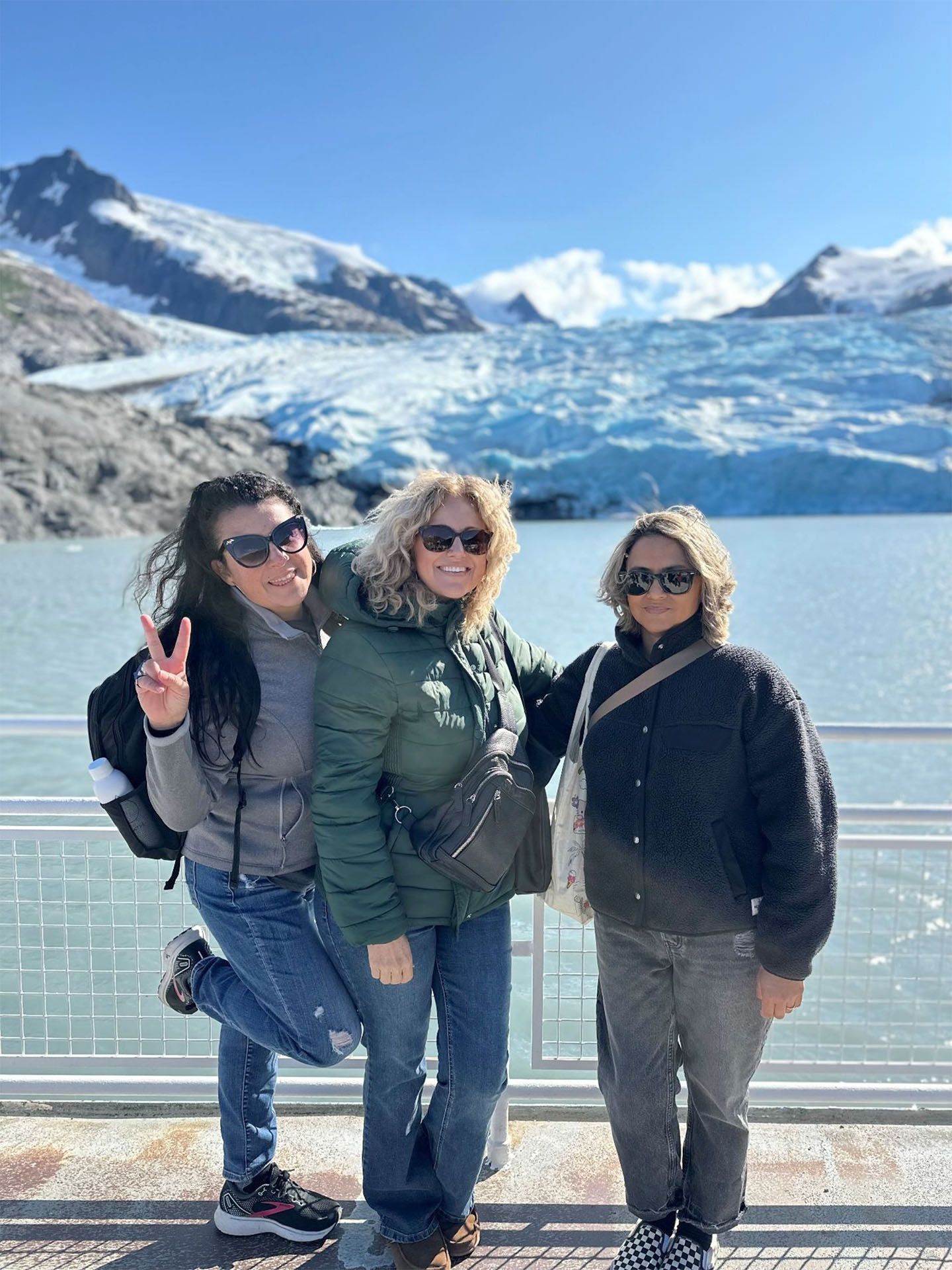 Three people pose in front of a glacier and water, wearing jackets and sunglasses
