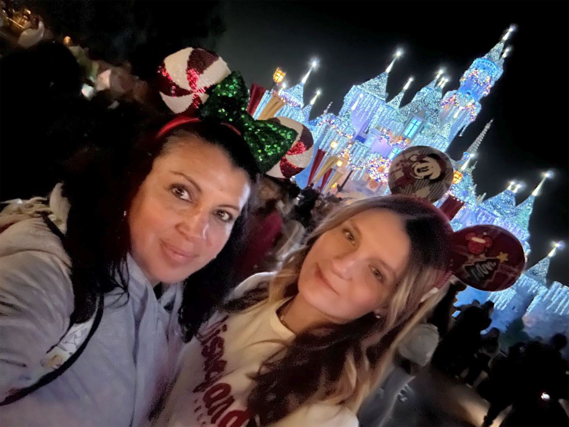 Two women smile, wearing Disney ears, in front of a brightly lit castle