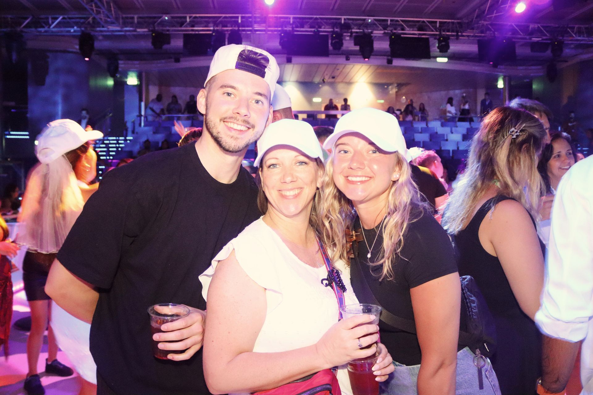 Three people smiling at a club, holding drinks. They wear white caps and dark clothes. Purple lighting.