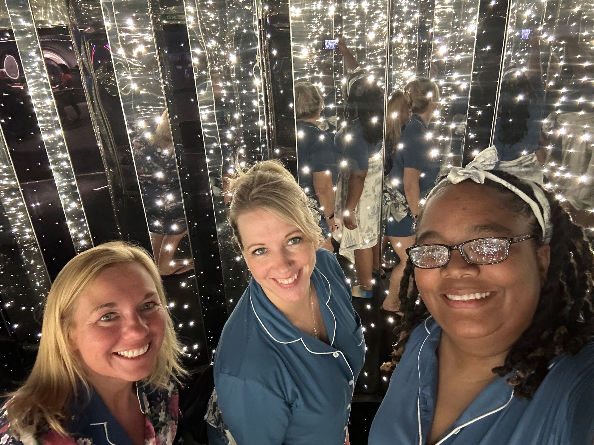 Three women in pajamas smile at the camera in a mirrored room with twinkling lights.