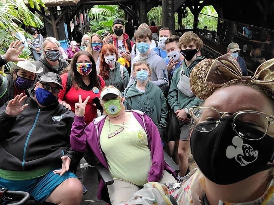 Group of people at a theme park wearing masks, some in wheelchairs, smiling.