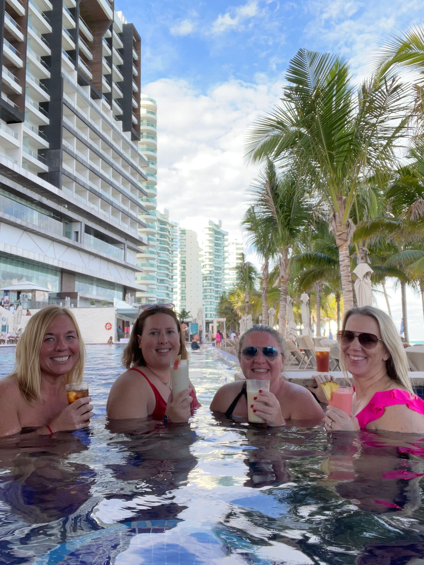 Four women in a pool, drinks in hand, smiling at the camera, tropical setting with high-rise buildings.
