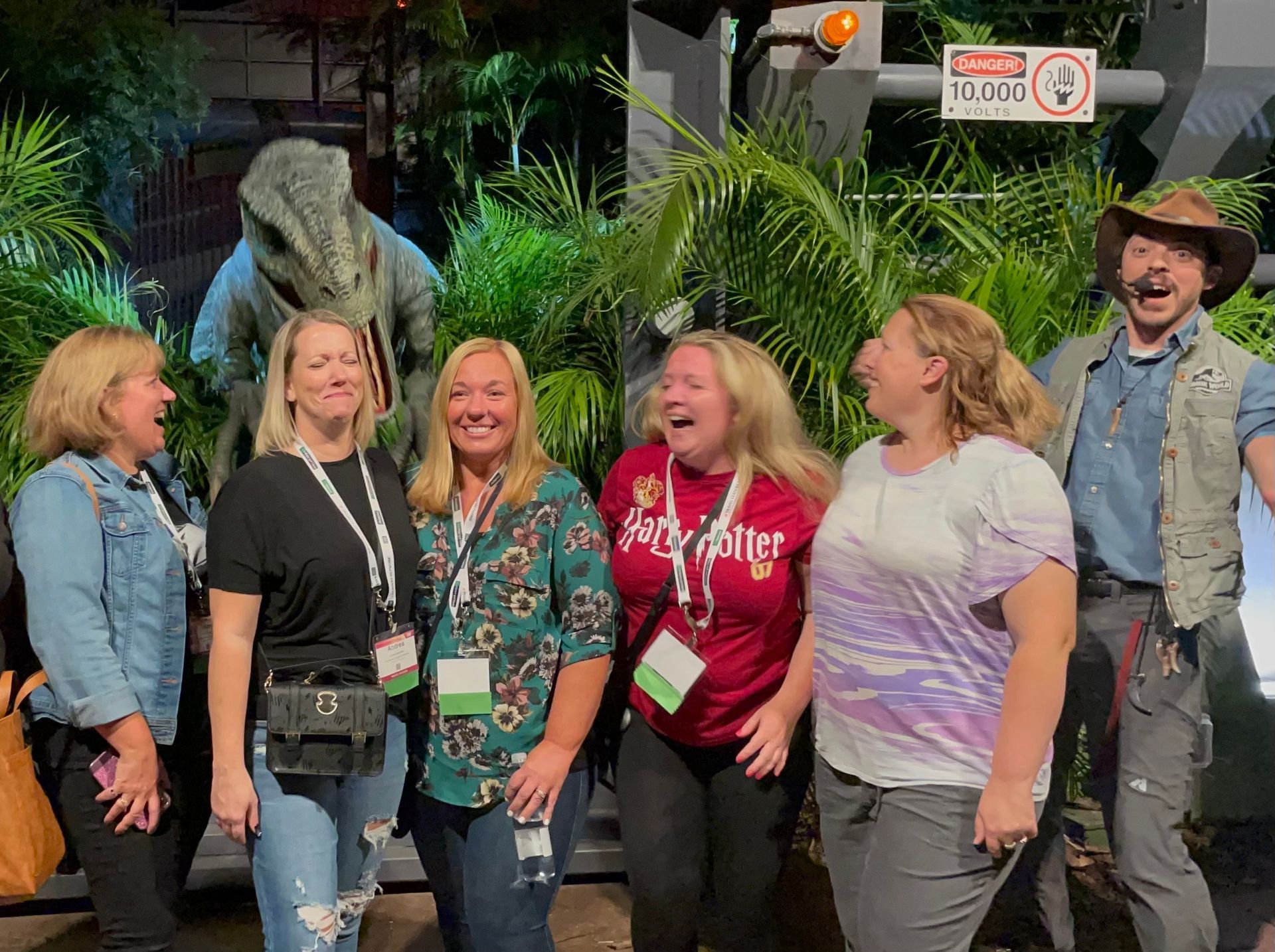 Group of women with a dinosaur and a park ranger, laughing, near tropical plants.