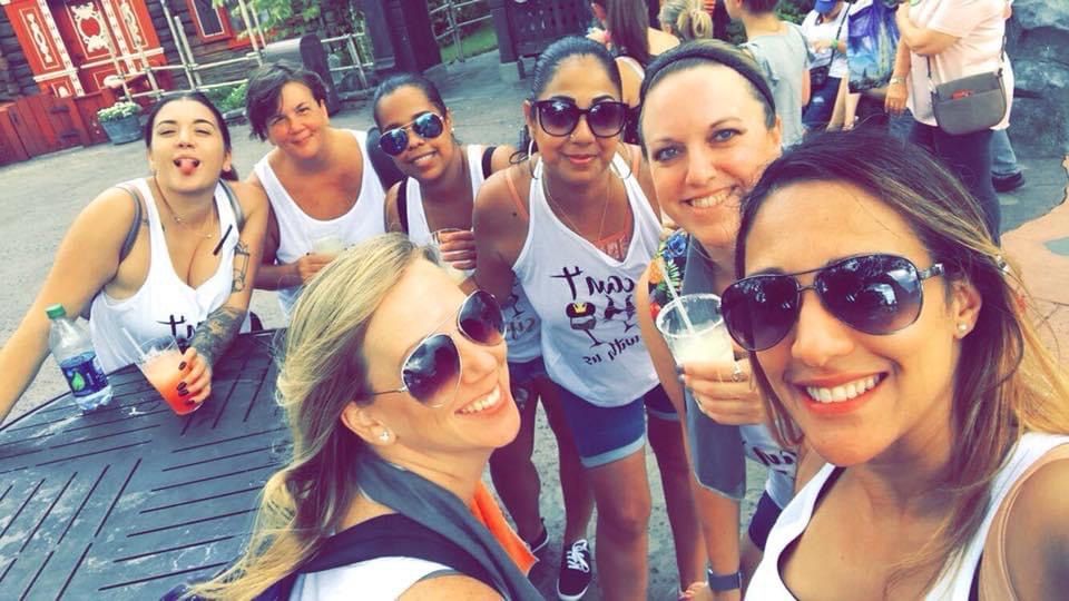 Group of women in sunglasses smiling, posing for a selfie outdoors. They wear white tank tops and hold drinks.