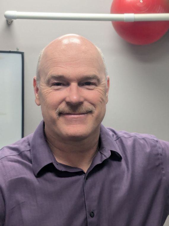 A bald person with a mustache smiling while wearing a purple collared shirt in front of a neutral wall and red ball.