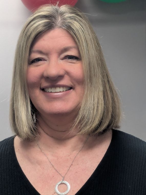 Smiling person with shoulder-length highlighted hair and a silver necklace, wearing a black top against a grey background.