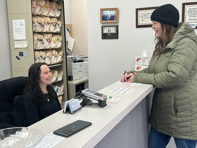 Two people interact at an office front desk; one seated at a computer, the other standing across the counter.