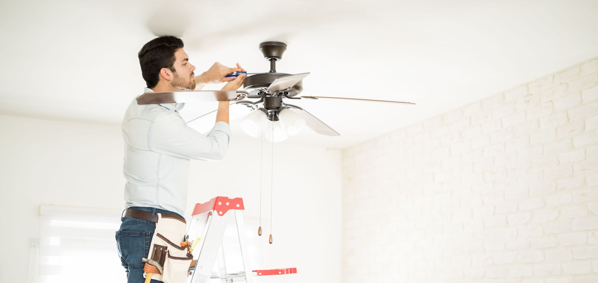 A person wearing a tool belt stands on a ladder, installing a dark-colored ceiling fan in a white room.