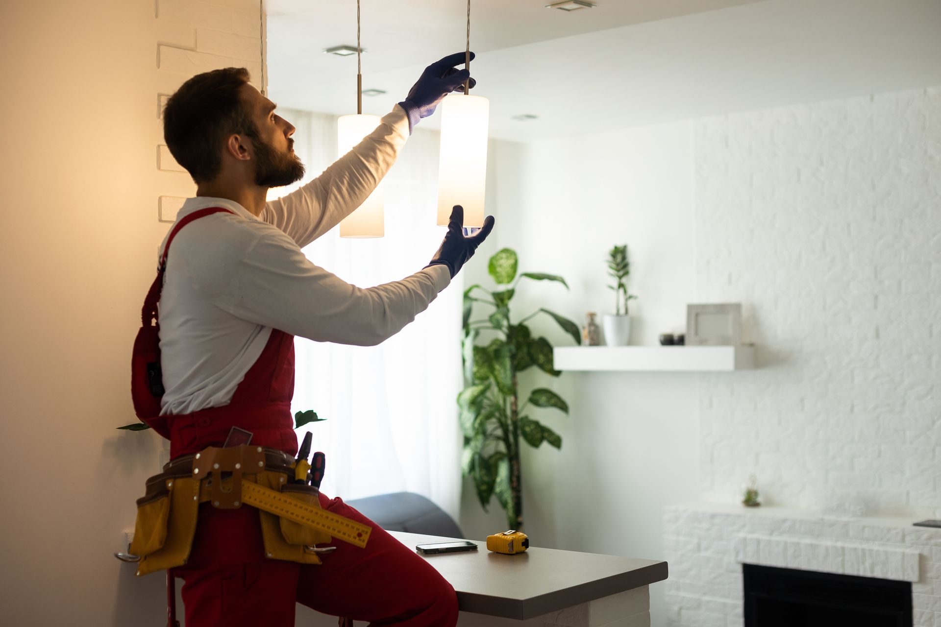 A person in red coveralls and a tool belt installs a cylindrical pendant light in a modern home.