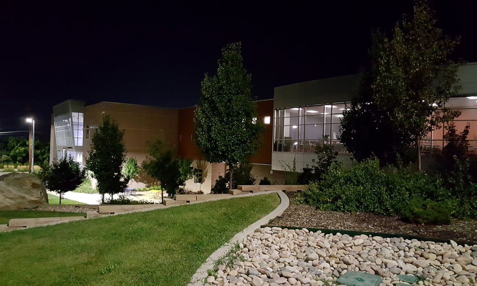 A modern building with large windows illuminated at night, viewed from a landscaped lawn with trees and a stone path.