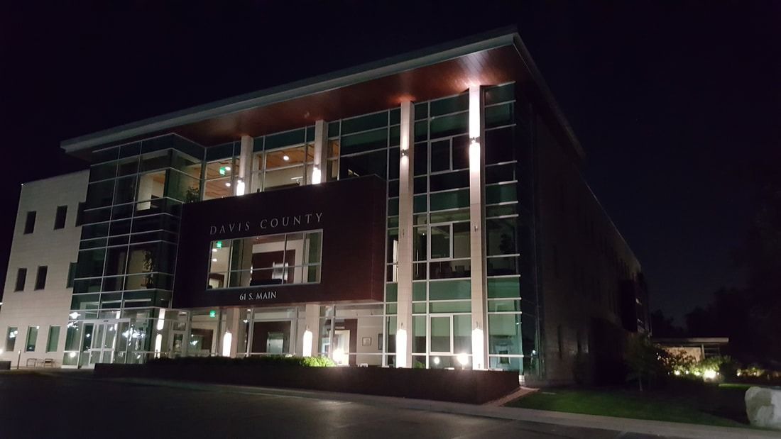 A modern office building at night with lit floor-to-ceiling glass windows and a prominent, dark-paneled entrance.
