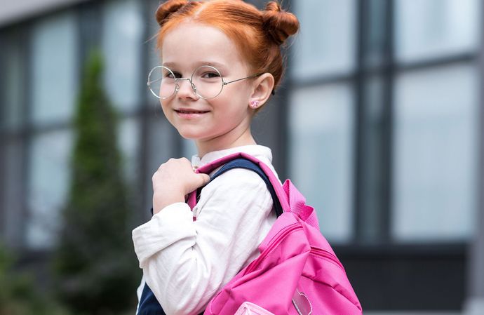 A little girl with red hair is wearing glasses and carrying a pink backpack.