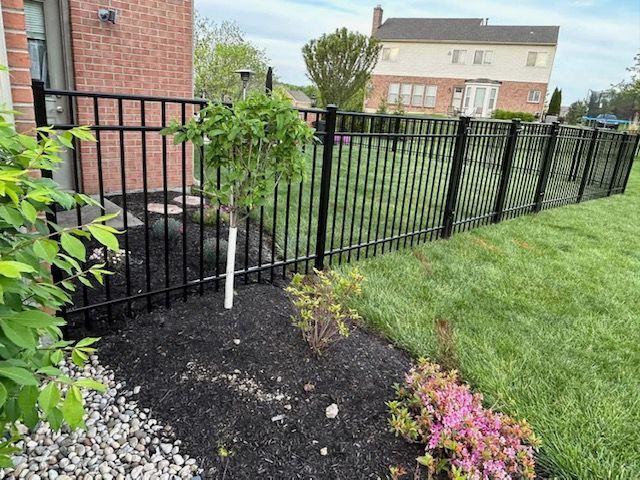 Black metal fence surrounding a yard with a brick house and landscaped flower beds.