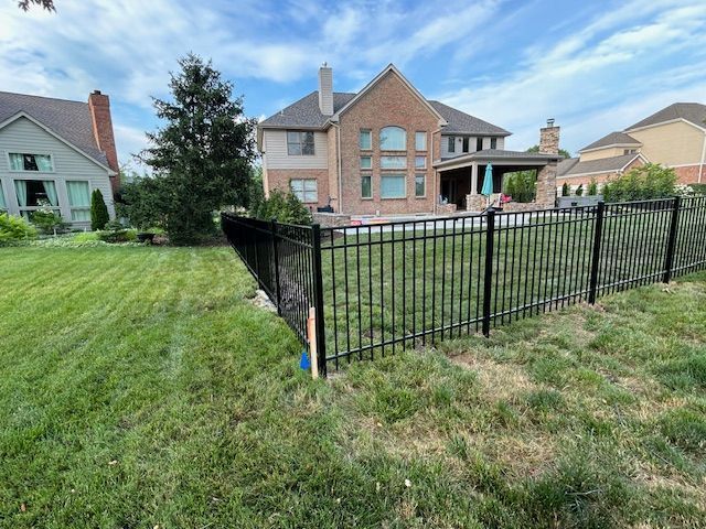 Black metal fence in a green yard, surrounding a pool near a large brick house under a blue sky.