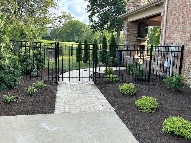 Black metal fence with gate along brick path, bordering mulch and landscaping near a house.