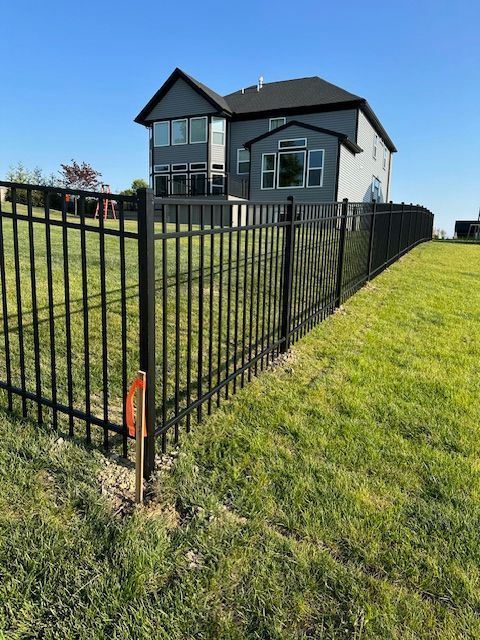 Black metal fence surrounds a grassy yard, with a two-story gray house in the background under a blue sky.