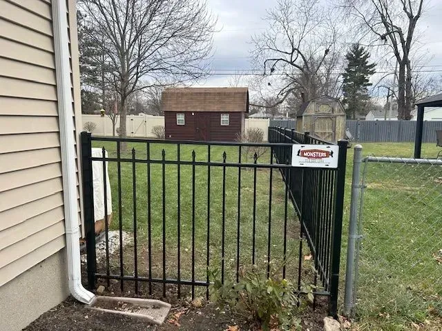 Black metal fence section near a house and backyard. Includes a brown shed and trees.