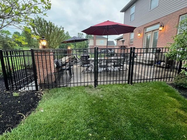 Black iron fence encloses a patio with outdoor furniture, umbrellas, and a brick feature, on a lawn.