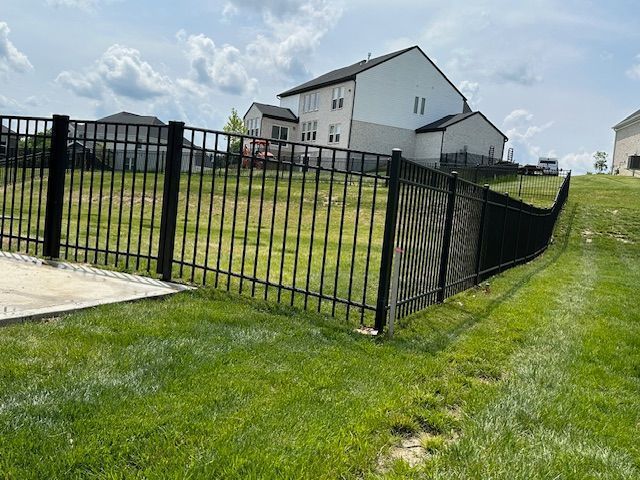 Black metal fence enclosing a grassy yard, sloping downward toward a house on a sunny day.