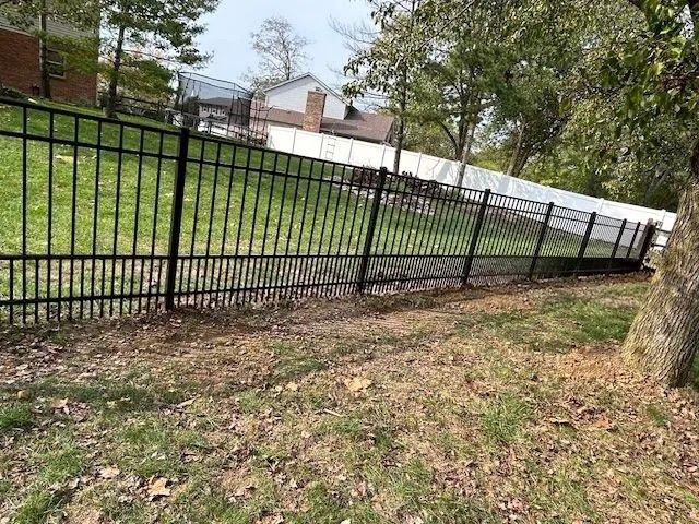 Black metal fence on a grassy hill, leading towards a white fence in the background, near trees and houses.